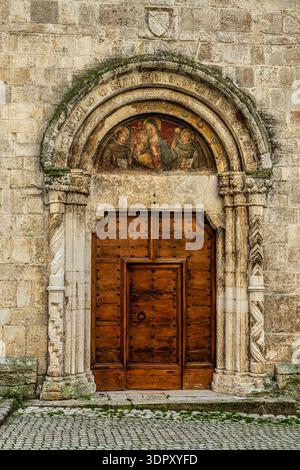 Historisches Steinportal einer italienischen Kirche mit einer alten Holztür, kunstvollen Schnitzereien und einem antiken religiösen Fresko in der Lünette. Pescina, Abruzzen Stockfoto