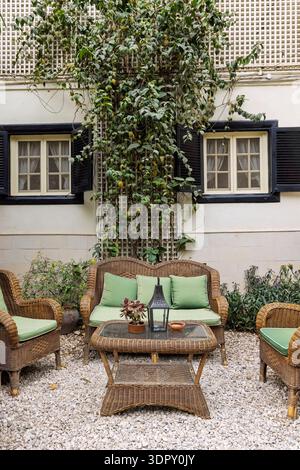 Terrassenmöbel aus Korb mit grünen Kissen, die auf einem kiesigen Innenhof unter Kletterstöcken und Fensterläden angeordnet sind, schaffen eine gemütliche, entspannte Garde Stockfoto