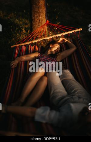 Frau, die sich in einer gestreiften Hängematte bei Sonnenuntergang mit einem Freund in der Nähe in einem Garten entspannt Stockfoto