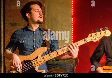 Babsea CD-Präsentation männlicher Musiker, der E-Bass-Gitarre auf der Bühne in einem kleinen Veranstaltungsort spielt, warmes rotes Licht, Backsteinmauerhintergrund, Live-Konzert Stockfoto