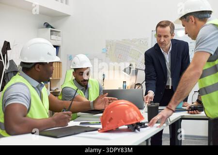 Kaukasischer Mann mittleren Alters im Anzug, der eine Gruppe junger Erwachsener, multiethnischer männlicher Vorarbeiter anweist, Sicherheitswesten und Helme trägt, am Bürotisch sitzt und Baupläne diskutiert Stockfoto