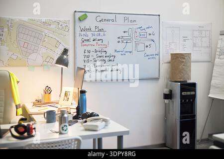 Büroarbeitsplatz mit Whiteboard mit Bauprojektplänen und Zeitplänen, Architekturzeichnungen an der Wand, Schreibtisch mit Computer und Kopfhörern, Wasserspender im Hintergrund Stockfoto