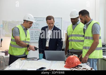 Kaukasischer Mann mittleren Alters im Anzug, der mit zwei Männern mittleren Alters in Sicherheitswesten und Schutzhüten und einem Schwarzen in Sicherheitsweste steht und Projektpläne im Büro mit Laptop bespricht Stockfoto