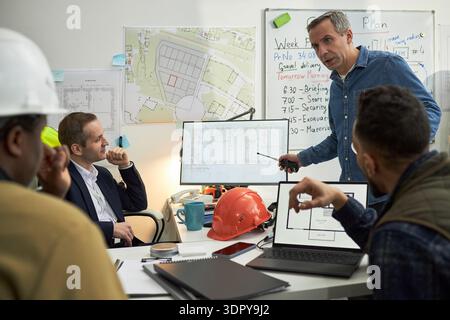 Kaukasischer Mann mittleren Alters stehend und mit Stift gespendet, während er ein Treffen mit verschiedenen Männern mittleren Alters im Amt führt und über Baupläne auf Monitoren diskutiert Stockfoto