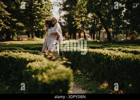 Zwei Frauen teilen sich einen verspielten Huckepack-Moment in einem sonnigen Park-Garten-Setting Stockfoto