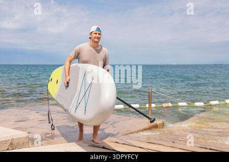 Ein junger Mann greift ein Paddleboard, während er an einem sonnigen Sommertag am Strand entlang spaziert. Er genießt Wassersport und einen aktiven Lebensstil. Stockfoto