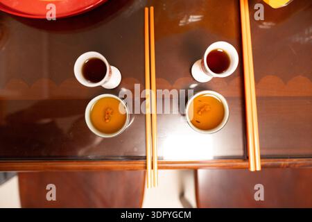 Blick von oben auf chinesischen Tee mit Essstäbchen auf Holztisch, dunkle und bernsteinfarbene Tees in Keramikbechern Stockfoto