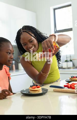 Afroamerikanische Mutter, die Sirup aus der Flasche über Pfannkuchen in der Küche gießt, die Tochter der Jugend beobachtet Stockfoto