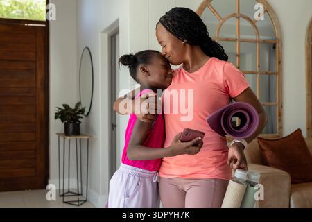 Afroamerikanische Mutter und Kind in Aktivkleidung umarmen, küssen im Eintritt, halten Yogamatte, Zelle Stockfoto