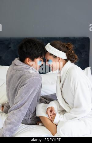 Verschiedene Paare, die auf dem Bett sitzen und sich gegenüberstehen, tragen weiche Bademäntel und blaue Flecken unter den Augen Stockfoto