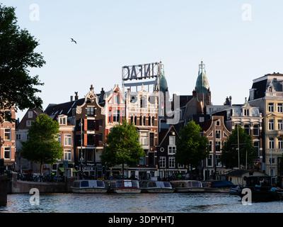 Amsterdam, Niederlande - 11. Mai 2025: Kanal mit Booten, die entlang des Damms neben einer Reihe historischer Gebäude mit Giebeldächern und Schildern in Niederländisch geparkt sind. Stockfoto
