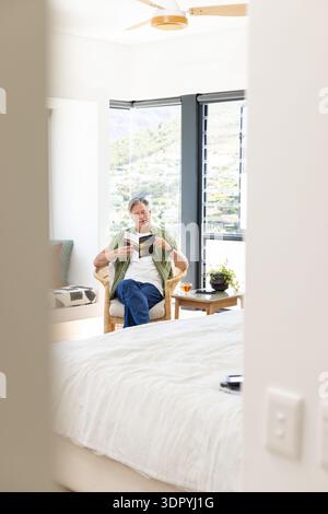 Mann mittleren Alters, der im Korbsessel sitzt und Taschenbuch im Schlafzimmer in der Nähe des Eingangs liest Stockfoto
