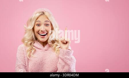 Frau mit blondem Haar lächelt und zeigt nach oben, während sie einen rosa Hoodie vor rosa Hintergrund trägt Stockfoto