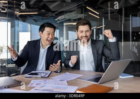 Zwei verschiedene Geschäftskollegen feiern einen Sieg, jubeln und lächeln über einem Laptop mit Finanzdokumenten auf einem modernen Schreibtisch während der täglichen Teamarbeit und des Erfolgs Stockfoto