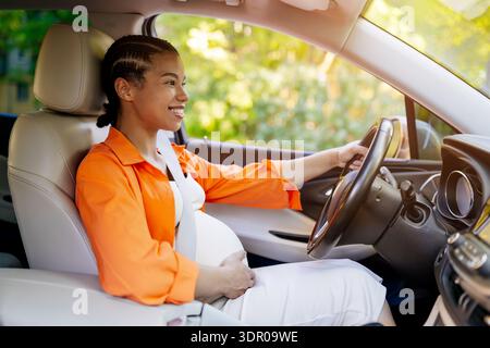 Junge schwarze schwangere Frau, die Auto fährt, lächelt und den Bauch hält Stockfoto