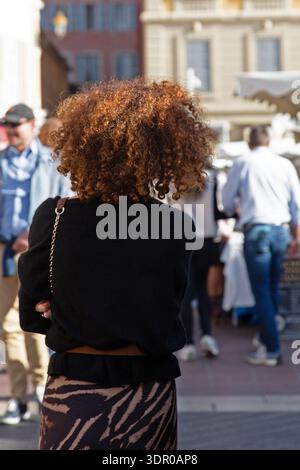Frau mit lockigem rotem Haar, die auf einem Markt im Freien in Nizza, Südfrankreich steht, mit Käufern und Ständen, die sanft verschwimmen. Stockfoto