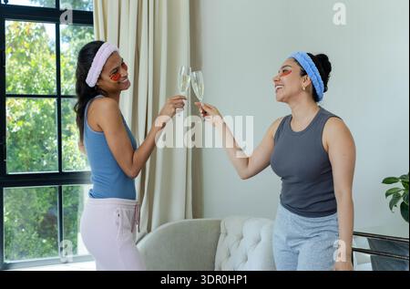 Zwei Freundinnen, die mit Sektflöten im Wohnzimmer tosten, tragen Stirnbänder und Gel-Patches Stockfoto