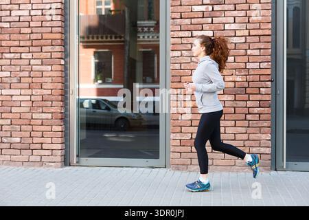 Die junge Frau läuft tagsüber in sportlicher Kleidung an einer Ziegelmauer in einer Stadt entlang Stockfoto