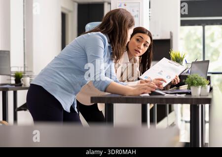 Weibliche Mitarbeiterinnen im Unternehmen, die sich mit Papieren auf den Schreibtisch stützen und gedruckte und Online-Unternehmensdaten vergleichen. Asiatische und kaukasische Geschäftsfrauen planen die Mittelzuweisung für die Projektentwicklung. Stockfoto