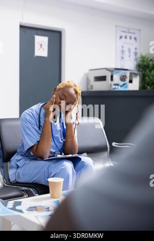 Die erschöpfte Krankenschwester ruht im Wartebereich des Krankenhauses, sichtbar müde und lehnt sich nach einem intensiven Arbeitstag nach vorne. Medizinisches Personal hat das Gesicht erschöpft, während es sich die Patientenakten auf der Zwischenablage ansieht. Stockfoto