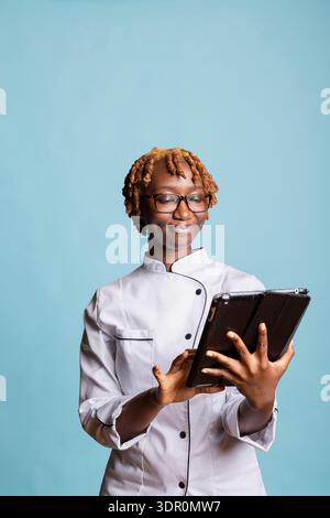 Weibliche Chefkochin studiert digitale Rezepte und überprüft sorgfältig die Schritte für die nächste kulinarische Kreation. Glückliche professionelle Köchin in Uniform verwendet Tablette, plant Gourmetgerichte und organisiert Zutaten. Stockfoto