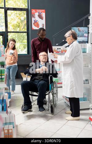 Leitender Apotheker, der Rezepte mit älteren Patienten im Rollstuhl überprüft und Ratschläge zur Sicherheit von Medikamenten und zur richtigen Dosierung bietet. Unterstützung einer zugänglichen und mitfühlenden pharmazeutischen Versorgung. Stockfoto