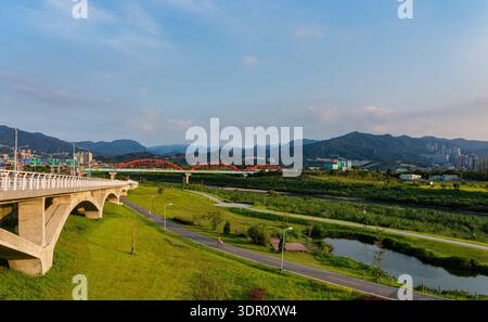 Die rote Bogenbrücke überspannt das Flusstal, umgeben von grünen Wegen und fernen Bergen. Stockfoto