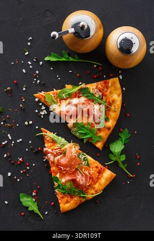Pizza-Scheiben mit Arugula und Prosciutto auf schwarzer Oberfläche mit Gewürzen Stockfoto