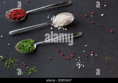 Anordnung von Gewürzen und Salz auf Löffeln auf dunkler Oberfläche mit verstreuten Inhaltsstoffen Stockfoto