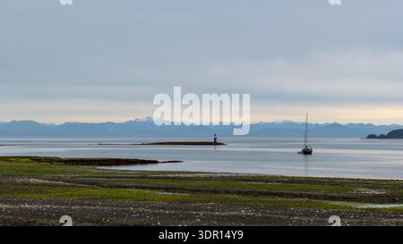 Port Hardy, BC, Kanada - 25. Juni 2024: Segelboot ankerte vor einem felsigen Strand von Port Hardy unter hohen Wolken mit Bergen in der Ferne. Stockfoto