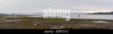 Port Hardy, BC, Kanada - 25. Juni 2024: Panoramablick auf ein Anker-Segelboot vor einem felsigen Strand von Port Hardy unter hohen Wolken und weit entfernten Bergen. Stockfoto