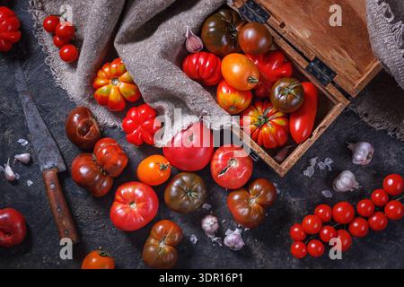 Viele verschiedene Arten, Formen und Größen von Tomaten in einer alten Holzkiste und auf einer dunklen Oberfläche, flach liegend, Draufsicht. Erntekonzept Stockfoto