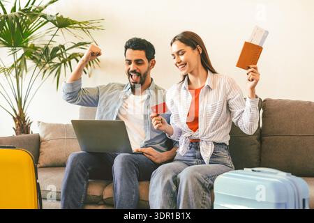 Aufgeregte Touristen paaren sich, die ein Hotelzimmer online buchen, mit Laptop und Kreditkarte, auf dem Sofa mit Koffern sitzen Stockfoto