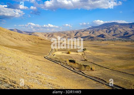 Eine gewundene Feldstraße führt durch ein ausgedehntes, trockenes Tal, umgeben von sanften braunen Bergen unter einem hellblauen Himmel in der Nähe von Van, Türkei Stockfoto