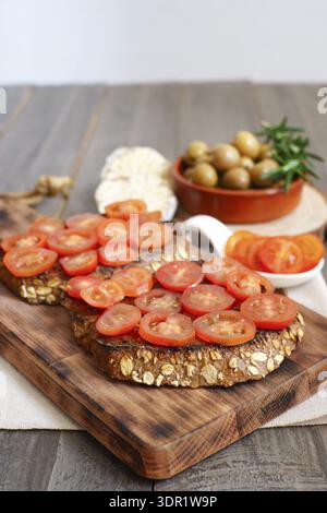 Zubereitung gesunder mediterraner Tapas mit in Scheiben geschnittenen Kirschtomaten auf geröstetem Brot und einer Seite grüner Oliven Stockfoto