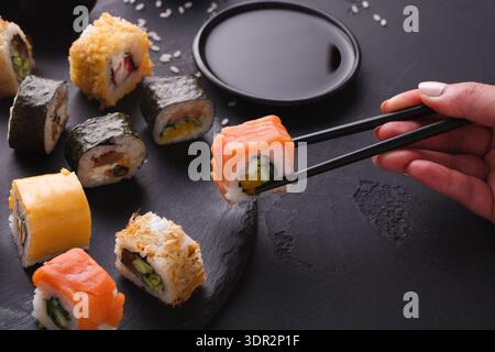 Sammlung von Sushi Rolls auf einem schwarzen Teller mit Sauce und Stäbchen in der Hand Stockfoto