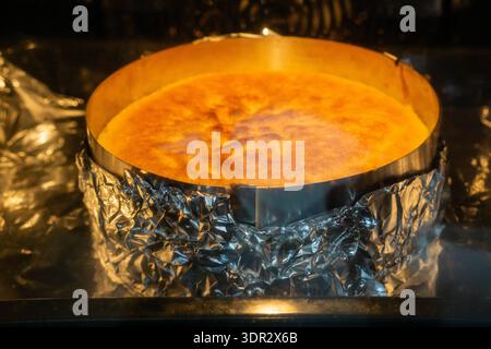 Ein runder Biskuitkuchen wird in einem Ofen in einem mit Folie ausgelegten Metallring gebacken. Die Oberfläche ist goldbraun geworden und zeigt eine perfekt aufgestiegene Kruste. Stockfoto