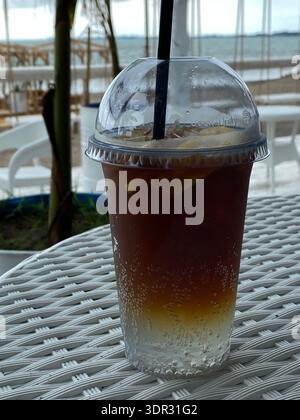 Eistee mit Zitrone in Plastikbecher mit Strohhalm auf einem Café-Tisch im Freien in der Nähe eines verschwommenen Meeres Hintergrund. Mobiles Foto Stockfoto