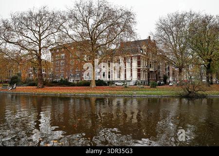 Herbstbäume und Backsteingebäude säumen den Kanal der Stadhouderskade und spiegeln sich an einem bewölkten Tag im Wasser in Amsterdam, Niederlande. Stockfoto