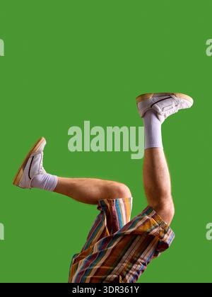 Männliche Beine in weißen Turnschuhen und gestreiften Shorts verspielte Pose auf grünem Hintergrund. Stockfoto
