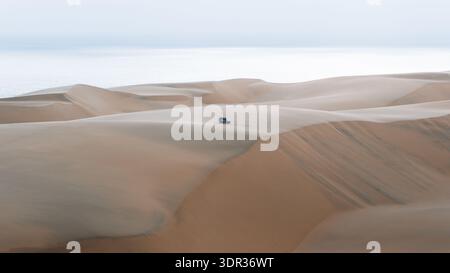 Der Blick auf ein einsames Auto fährt durch die hügeligen Dünen unter einem hellen Himmel und wirft Schatten über die sandigen Weiten, eine ruhige Wüstenlandschaft, Walvis Bay, N Stockfoto