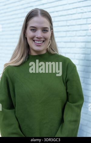 Erwachsene Frau lächelt in der Kamera an der weißen Ziegelwand in olivgrünem Pullover, Reifen, blonden Haaren Stockfoto