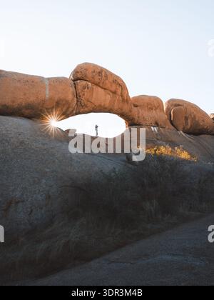 Blick auf das Sonnenlicht, das durch einen Natursteinbogen bricht, wo eine Silhouette in der Ferne steht, einen starken Kontrast von Schatten und Licht, Spitzkoppe, Stockfoto