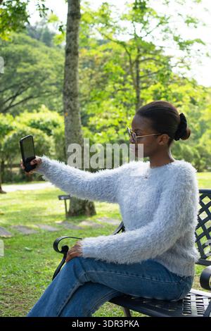 Stilvolle Frau, die Selfies mit Smartphone macht, während sie auf der Bank im grünen Park sitzt Stockfoto