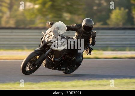 Ein Motorradfahrer in schwarzer Schutzausrüstung, der ein graues Abenteuer-Sportmotorrad während eines Rennkurses bei Sonnenuntergang in eine Kurve lenkt. Stockfoto