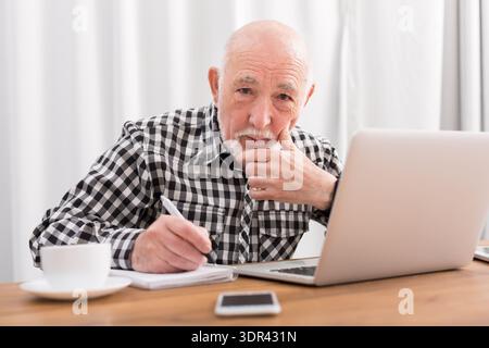 Ein älterer Mann sitzt an einem Tisch mit Laptop, Notizblock und Tasse Kaffee in einem hellen Raum Stockfoto