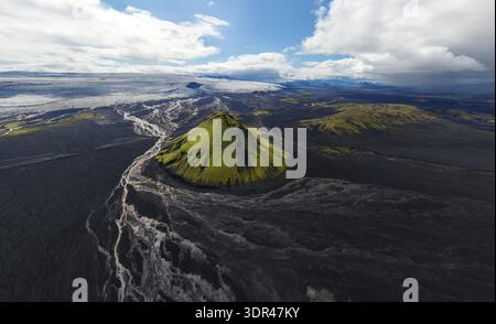 Aus der Vogelperspektive auf einen markanten grünen Hügel inmitten der krassen schwarzen Vulkanlandschaft, unter einem dynamischen Himmel, Rangárþing, Landmannaleið ytra, Island. Stockfoto
