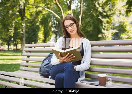 Genießen Sie ein Buch an einem sonnigen Tag in einem Park mit einem Kaffee in der Hand, während Sie auf einer Bank sitzen Stockfoto