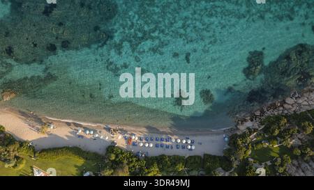 Aus der Vogelperspektive auf das pulsierende türkisfarbene Meer, das sanft den Sandstrand küsst, eingerahmt von zerklüfteten Felsen und grünem Grün, Porto Cervo, Sardinien, Italien. Stockfoto