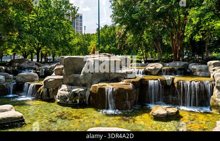 Centennial Olympic Park in Atlanta, Georgia. Malerischer, künstlicher Wasserfall fließt über Felsformationen, umgeben von grünen Bäumen und Stadtgebäuden unter blauem Himmel Stockfoto